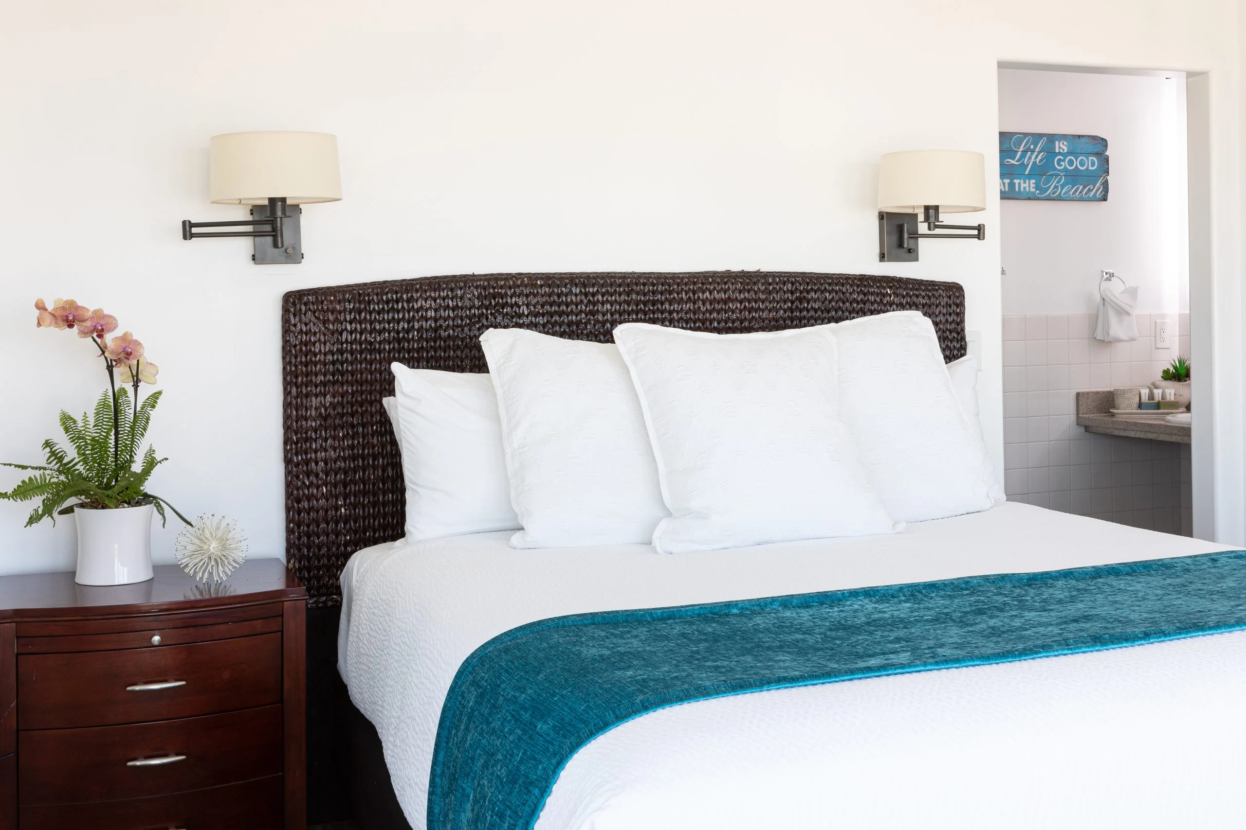 A neatly made bed with a wicker headboard, white bedding, and a teal throw, with a wooden nightstand and wall lamps beside it, and a bathroom doorway in the background.