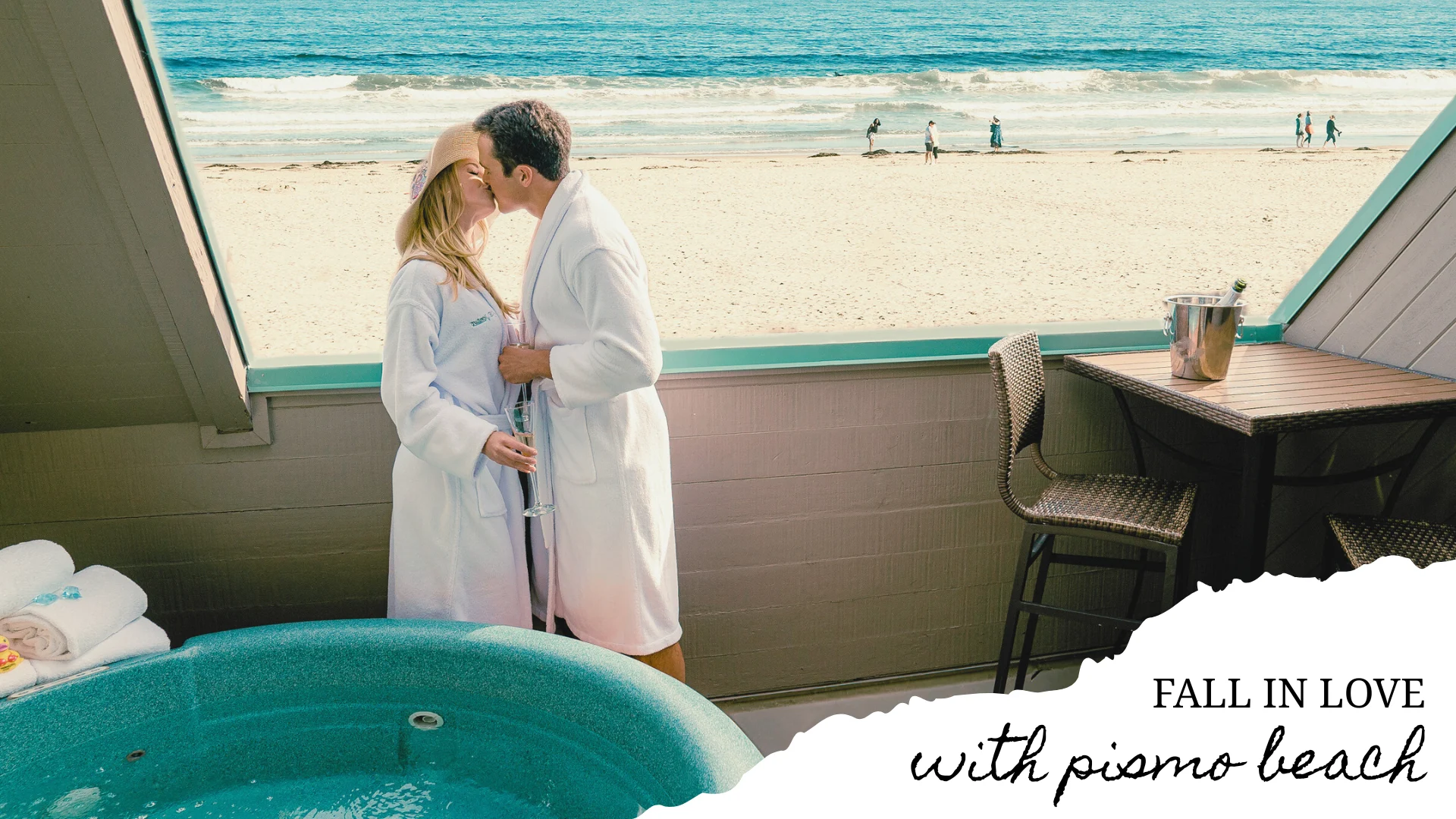A romantic couple in robes standing on a balcony with a hot tub, overlooking the beach and ocean.